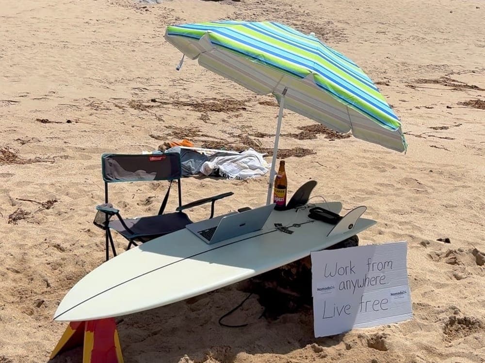 Surfbrett als Schreibtisch unter einem Sonnenschirm am Strand, daneben ein Schild mit der Aufschrift „Work from anywhere, live free“